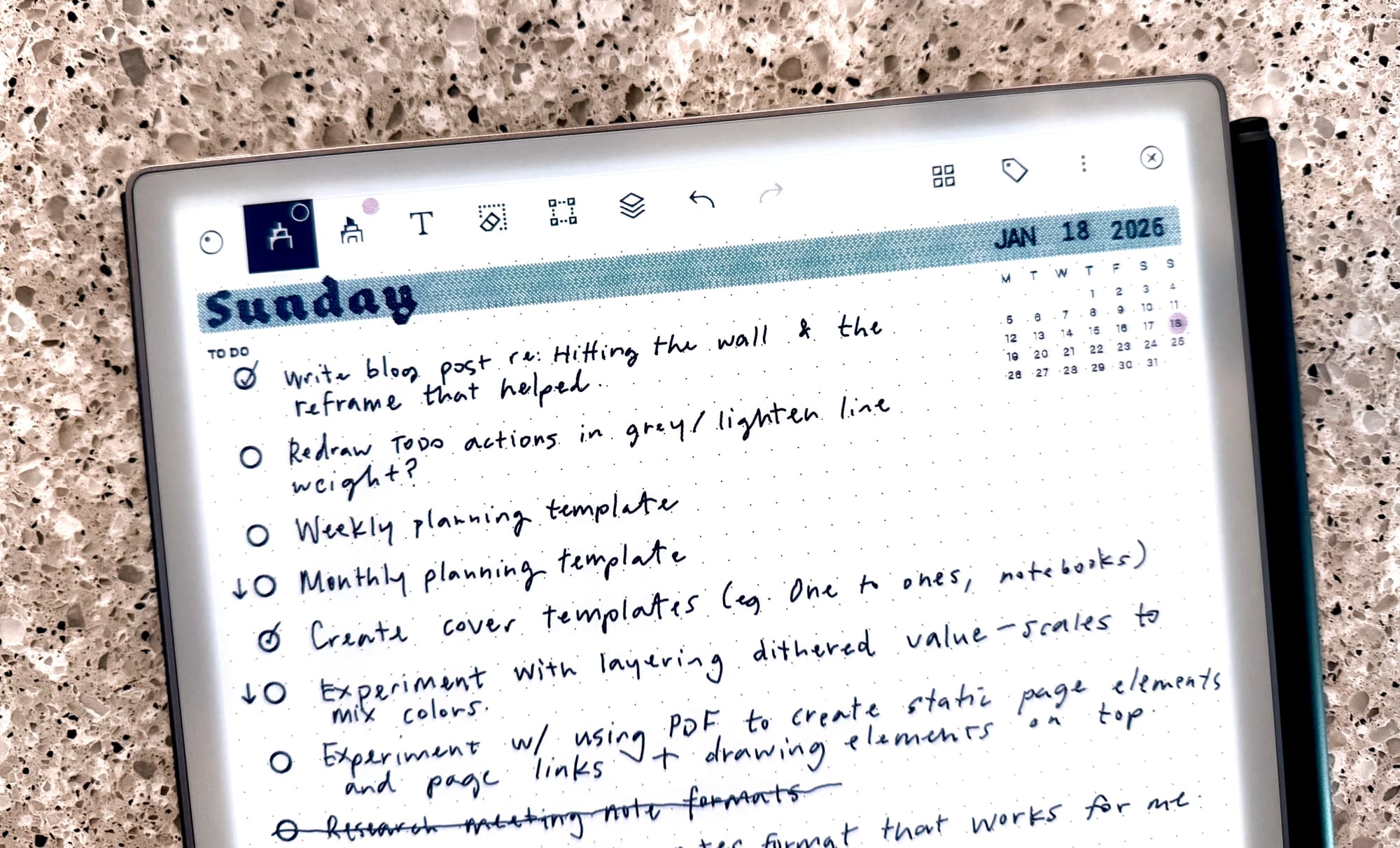 A digital tablet displaying a handwritten to-do list for Sunday, January 18, 2026, on a dotted grid interface. The tasks include writing a blog post, redrawing to-do actions, creating planning templates, experimenting with PDF layouts, and cover template design. The screen shows a mix of checked, unchecked, and in-progress items in black ink, laid out against a granite countertop background.