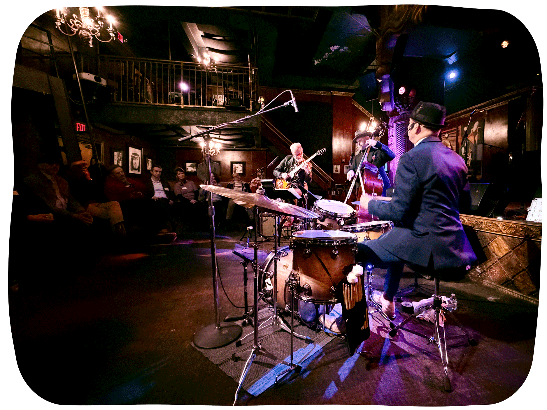 A jazz trio performs live in an intimate club with the drummer in the foreground and a guitarist and upright bassist playing under warm stage lights. Audience members sit close to the stage watching the musicians while chandeliers and a balcony add to the cozy lounge atmosphere. The image captures the energy of a small live music venue with instruments microphones and soft ambient lighting.
