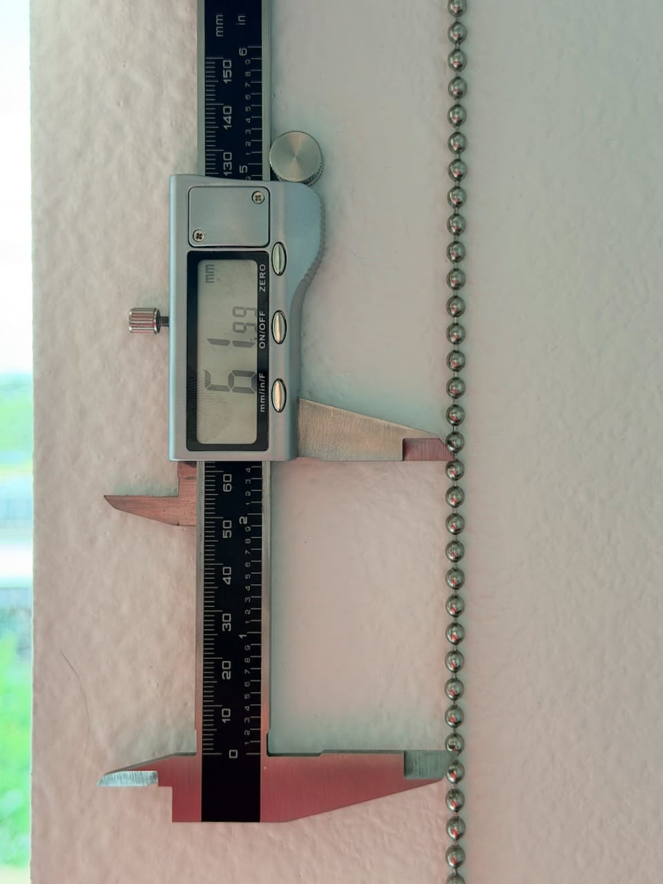 A digital caliper measuring the diameter of a metal ball chain against a wall.