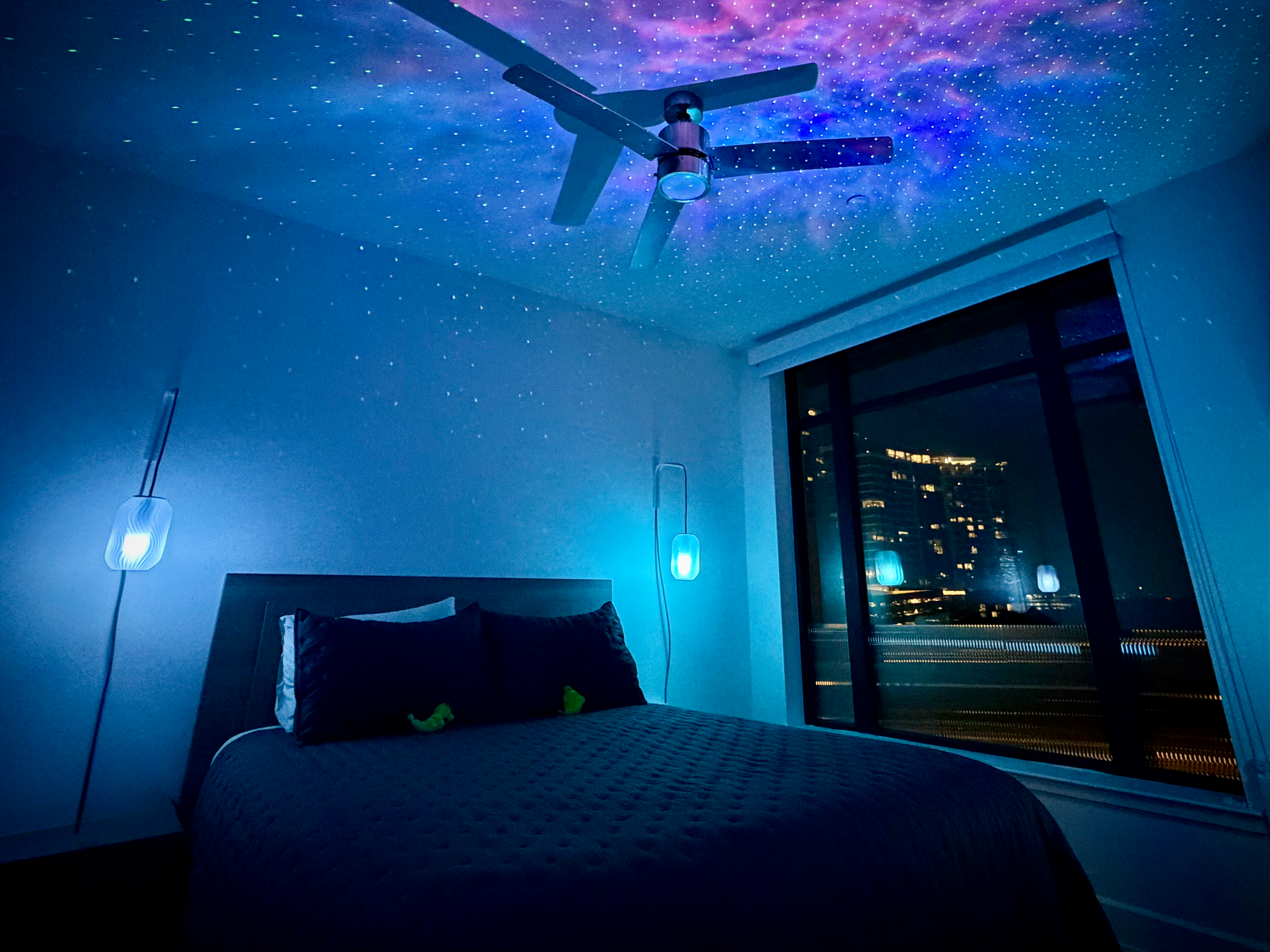 A dark bedroom at night with a star-projector ceiling glowing purple and blue, a ceiling fan, two illuminated wall sconces beside the bed, and a city skyline through the window.