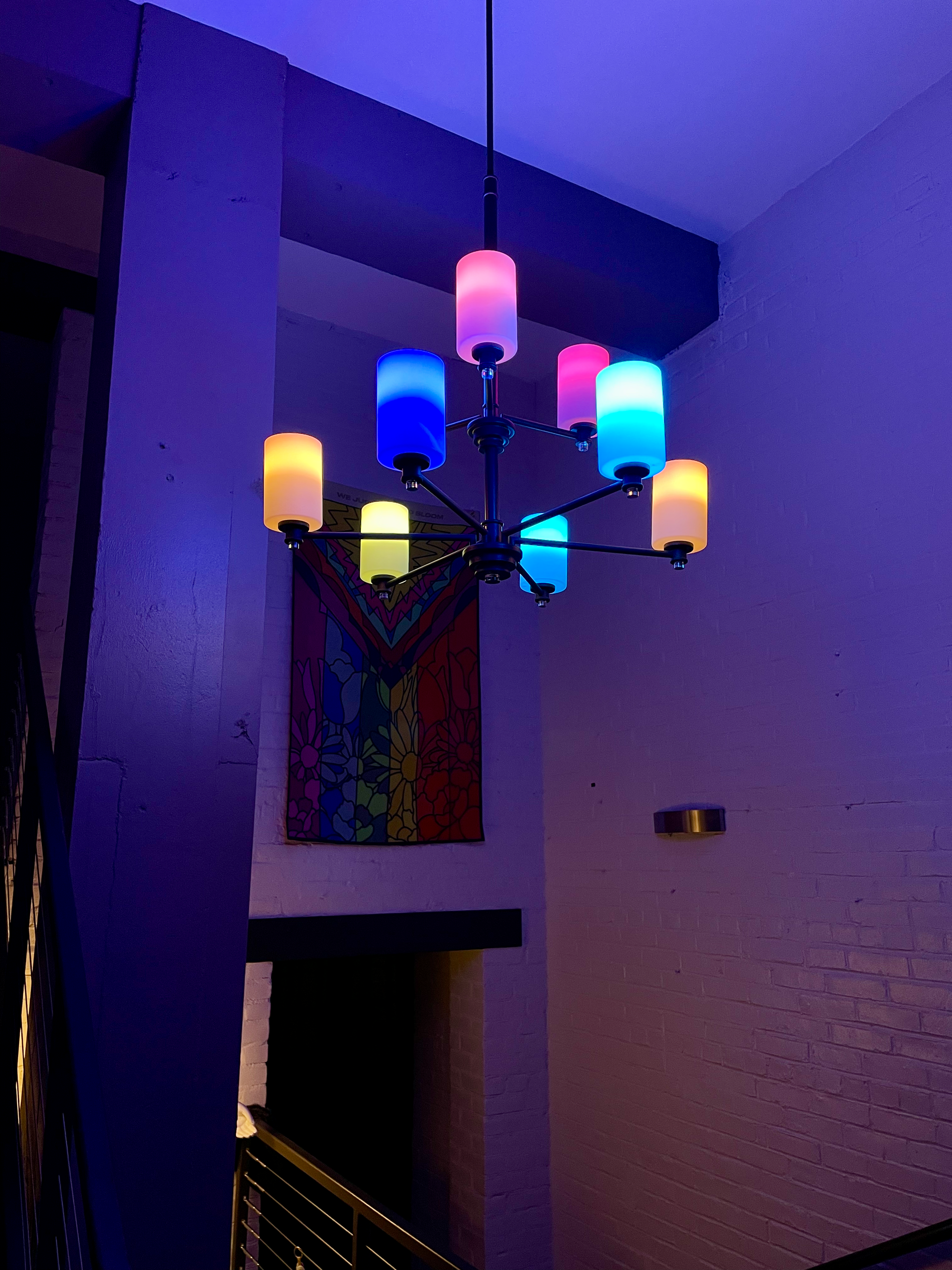 A multi-arm chandelier with colorful glass globe shades in pink, blue, magenta, cyan, yellow, and orange, hanging in a stairwell against an exposed brick wall with a rainbow tapestry below.