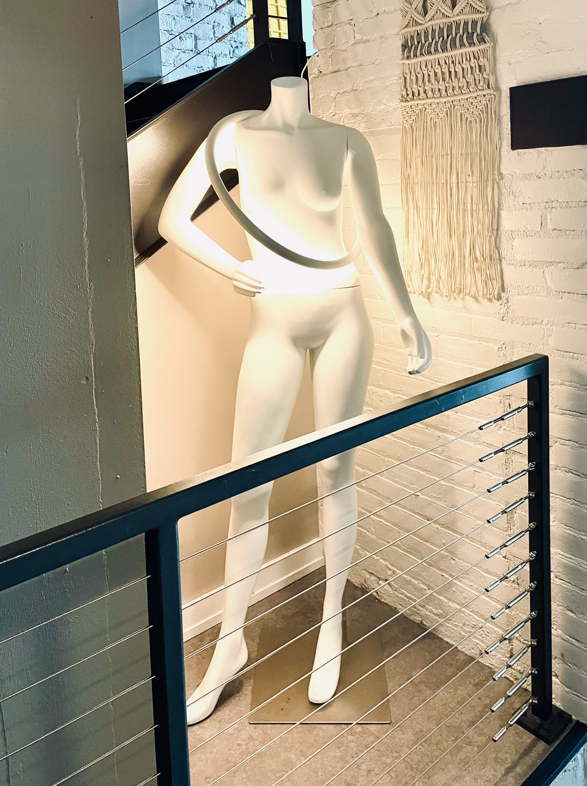 A headless mannequin torso draped with a glowing white ring light, standing against an exposed brick wall near a cable-railing staircase and a macrame wall hanging.