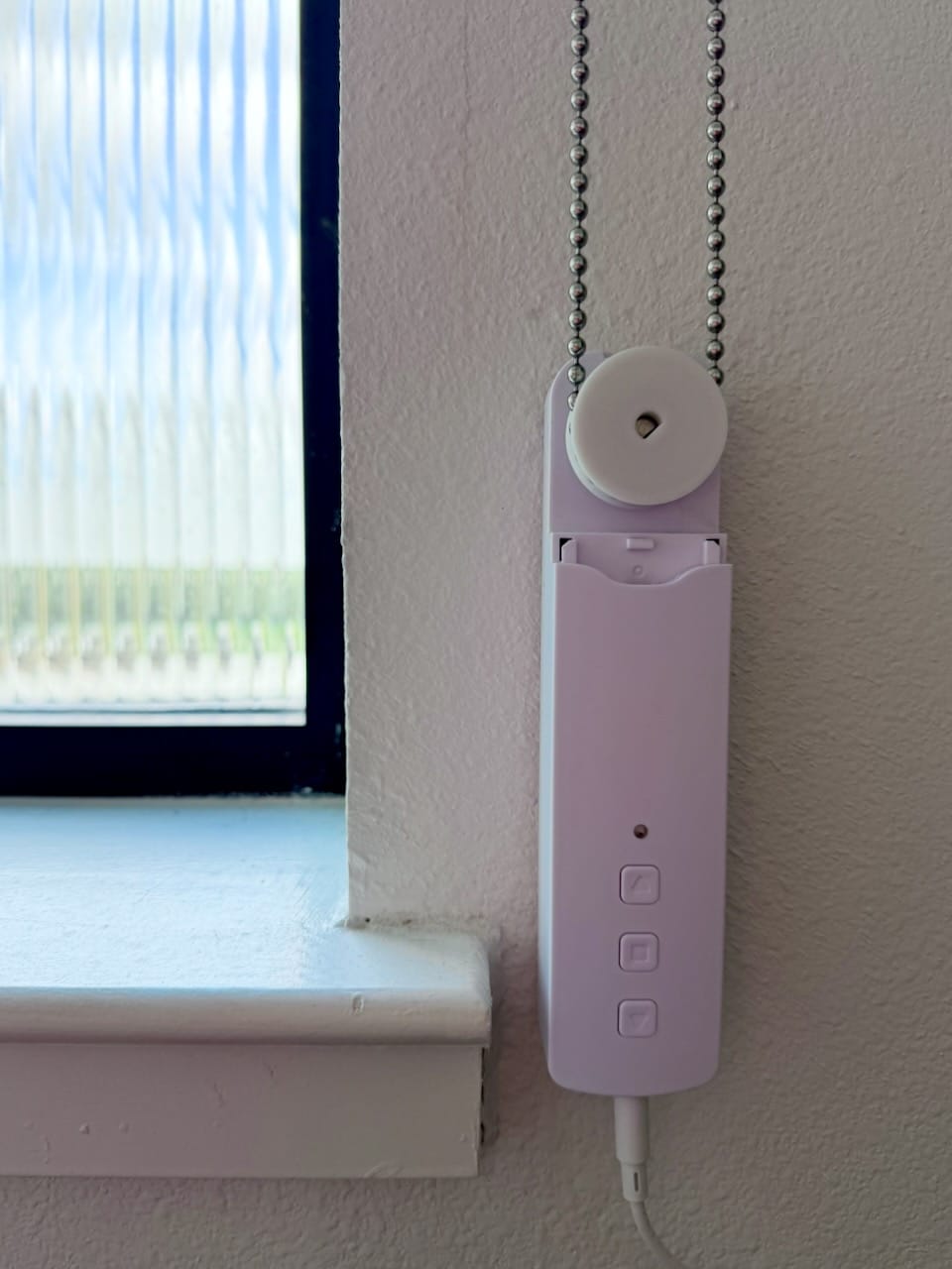 A custom white 3D-printed gear installed on a shade motor, gripping a metal bead chain, mounted on a wall beside a window.
