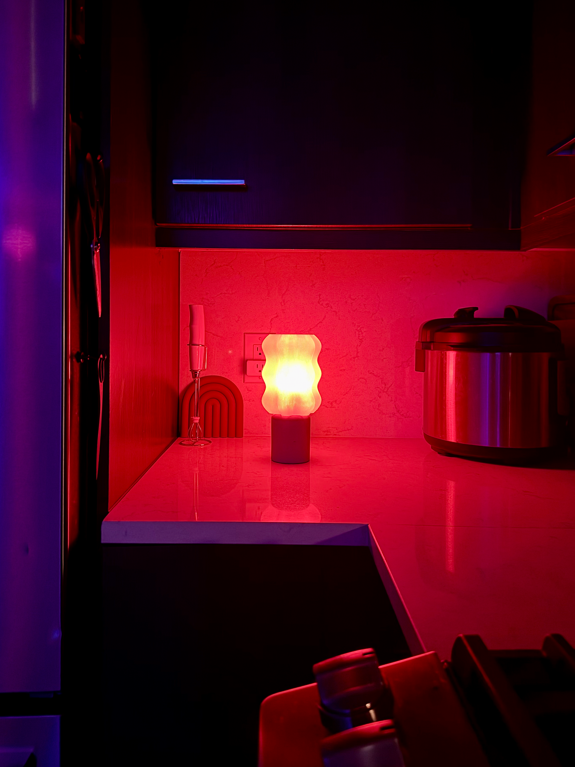 A warm red-orange glowing lamp on a dark kitchen counter at night, beside a small electric whisk and an arch-shaped hot pad, with an Instant Pot in the background.