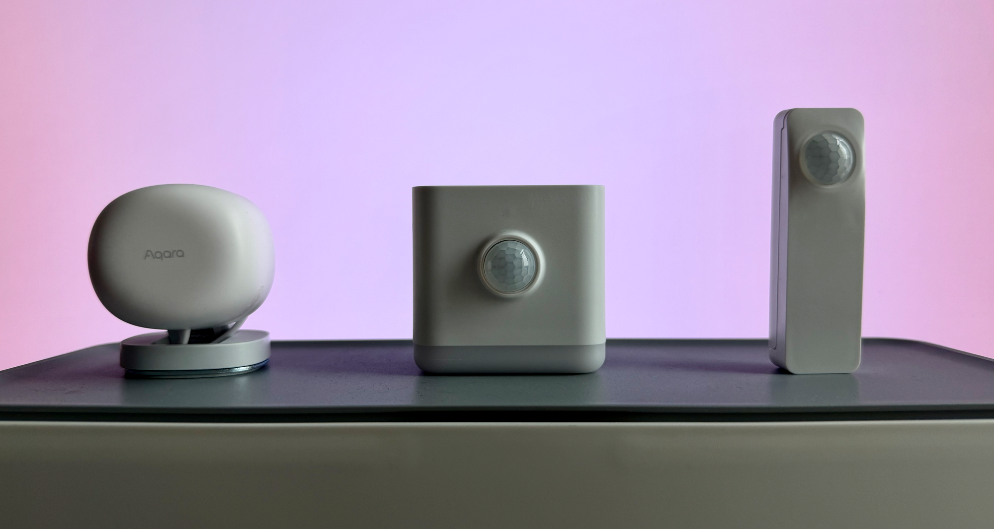 Three white smart home sensors lined up on a dark shelf against a pink-to-purple gradient background: a round Aqara presence sensor on the left, a square motion/lux sensor/nightlight in the middle, and a slim rectangular motion sensor on the right.