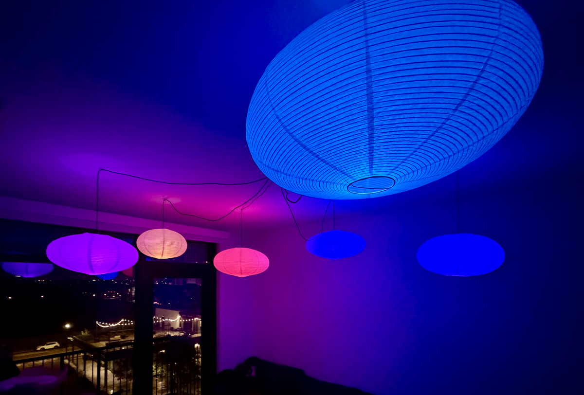 Six paper-lantern pendant lights glowing in blue, purple, and pink above a living room, with a city view visible through the window below at night.